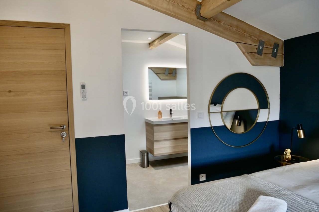 Chambre moderne avec poutres apparentes, mur bleu, lit double et vue sur une salle de bain attenante avec miroir éclairé.