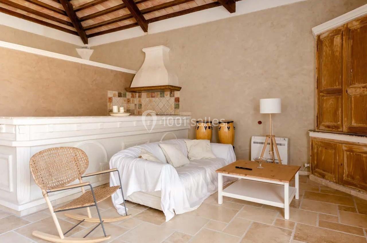 Salon chaleureux avec canapé blanc, fauteuil en osier, lampadaire, tambours et cheminée dans un décor rustique.