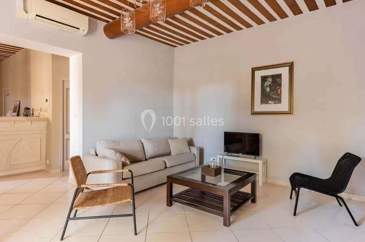 Salon lumineux avec canapé beige, table basse en verre, deux fauteuils, télévision et poutres apparentes au plafond.