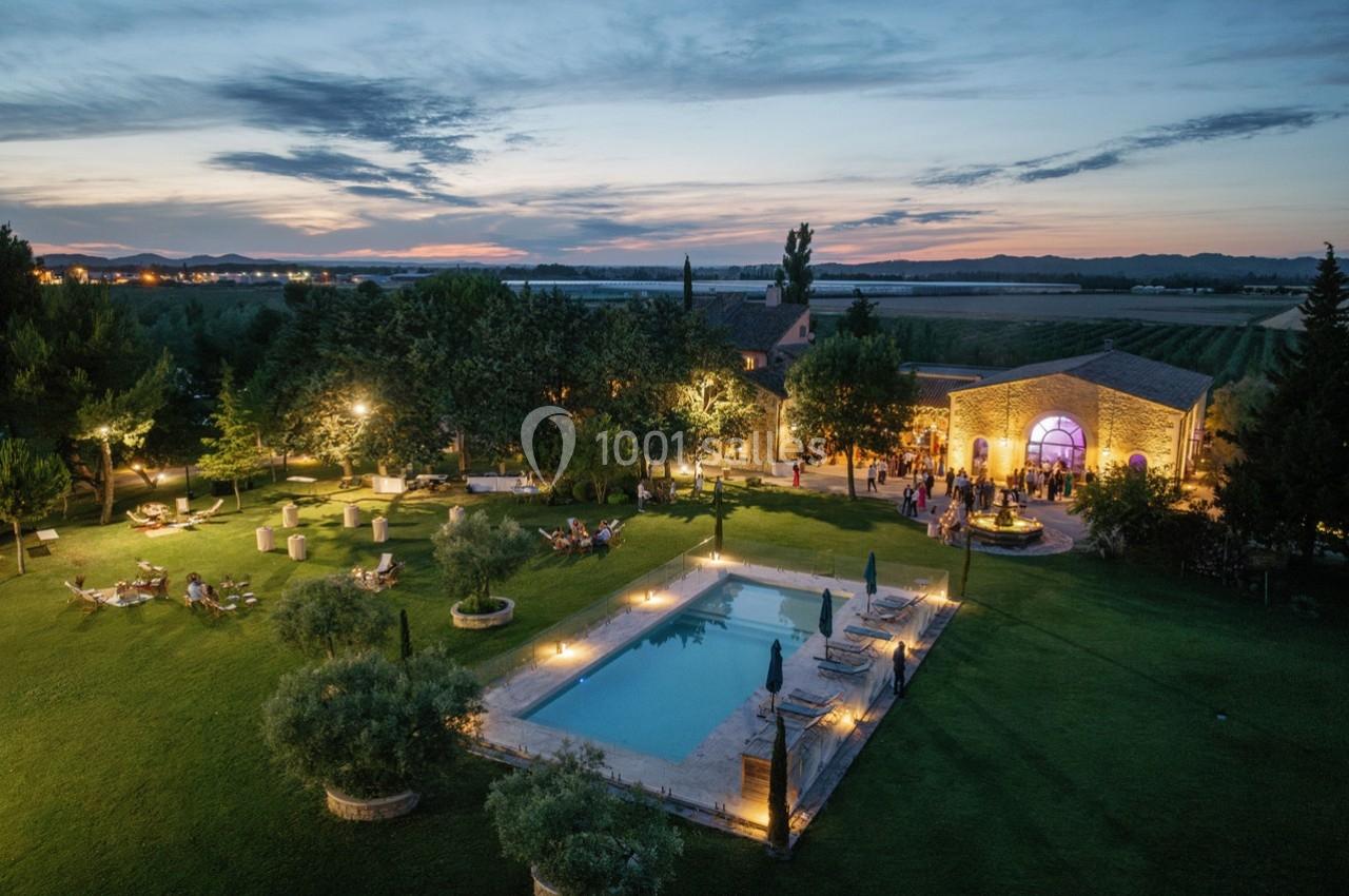 Vue aérienne d'une propriété avec piscine éclairée, jardin aménagé et bâtiment en pierre au crépuscule.