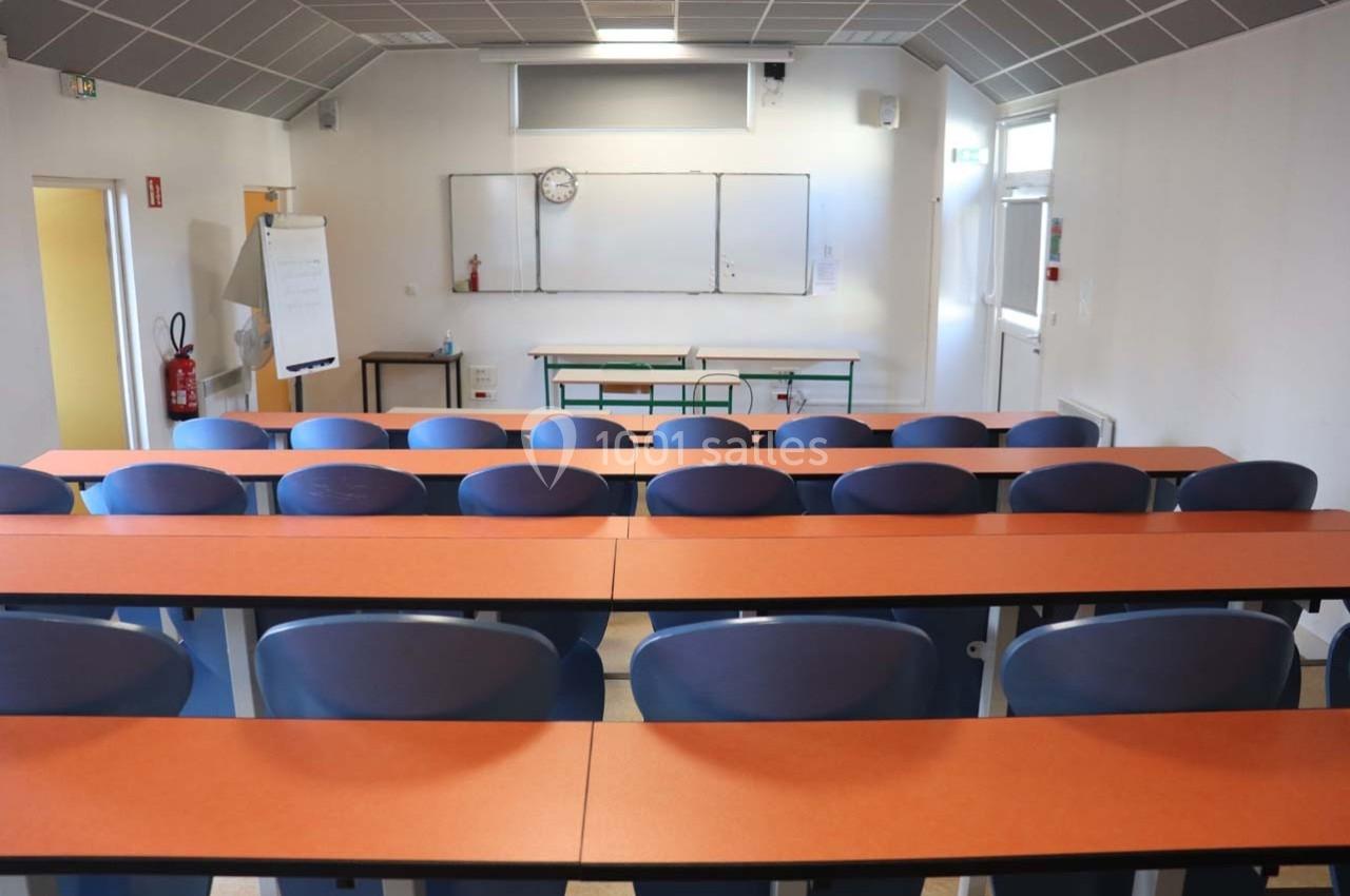 Salle de classe vide avec des rangées de tables et chaises, tableau blanc et horloge murale en arrière-plan.
