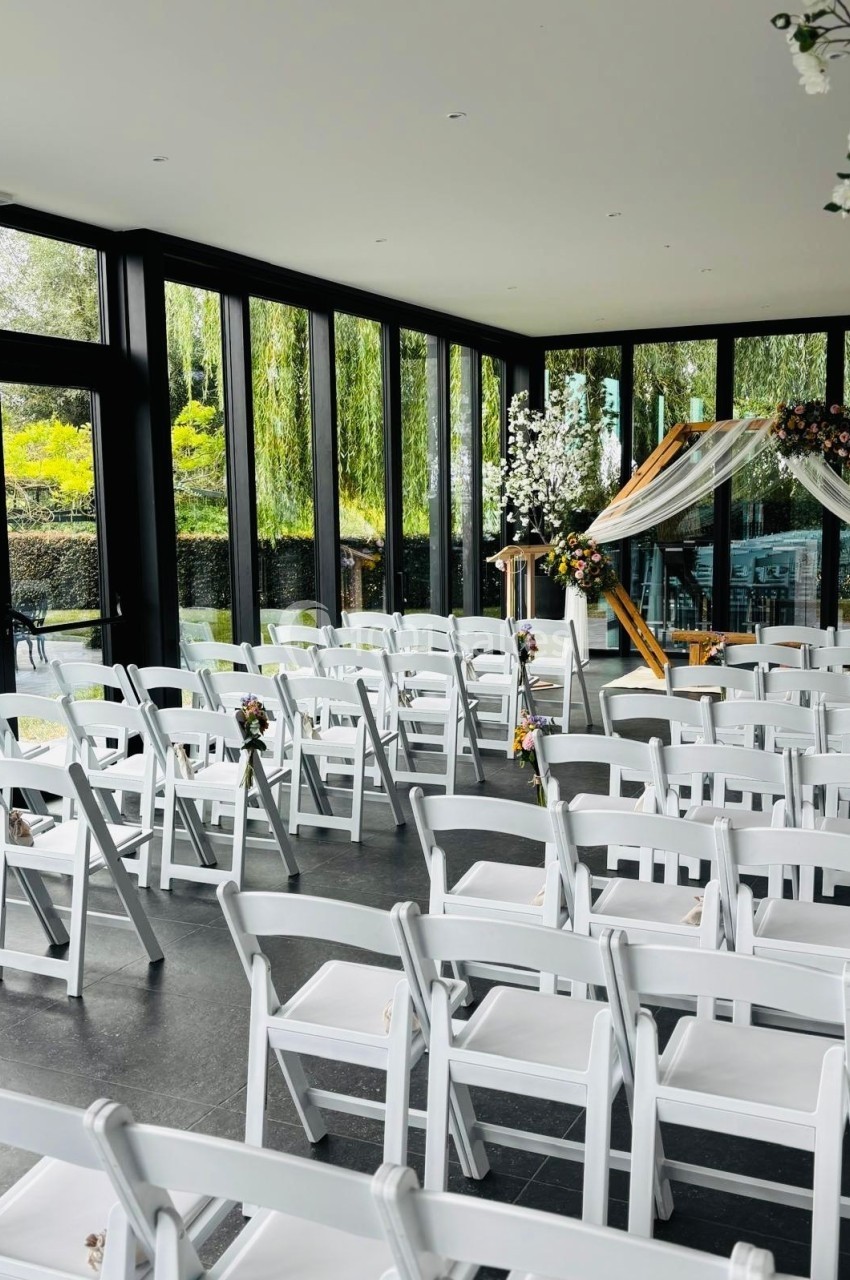 Chaises blanches alignées dans une salle vitrée avec arche décorée de fleurs pour une cérémonie.
