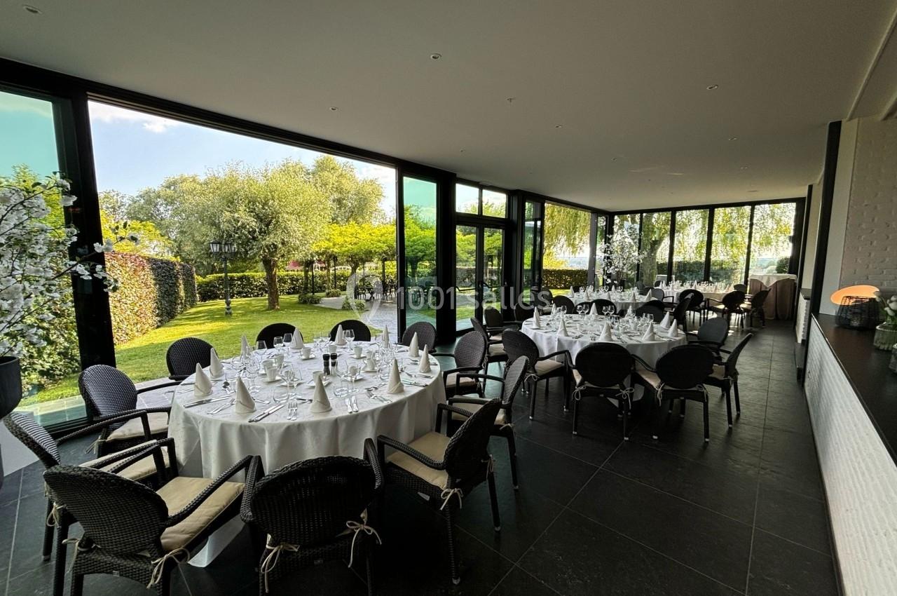 Salle lumineuse avec grandes baies vitrées donnant sur un jardin, tables rondes dressées pour un repas formel.