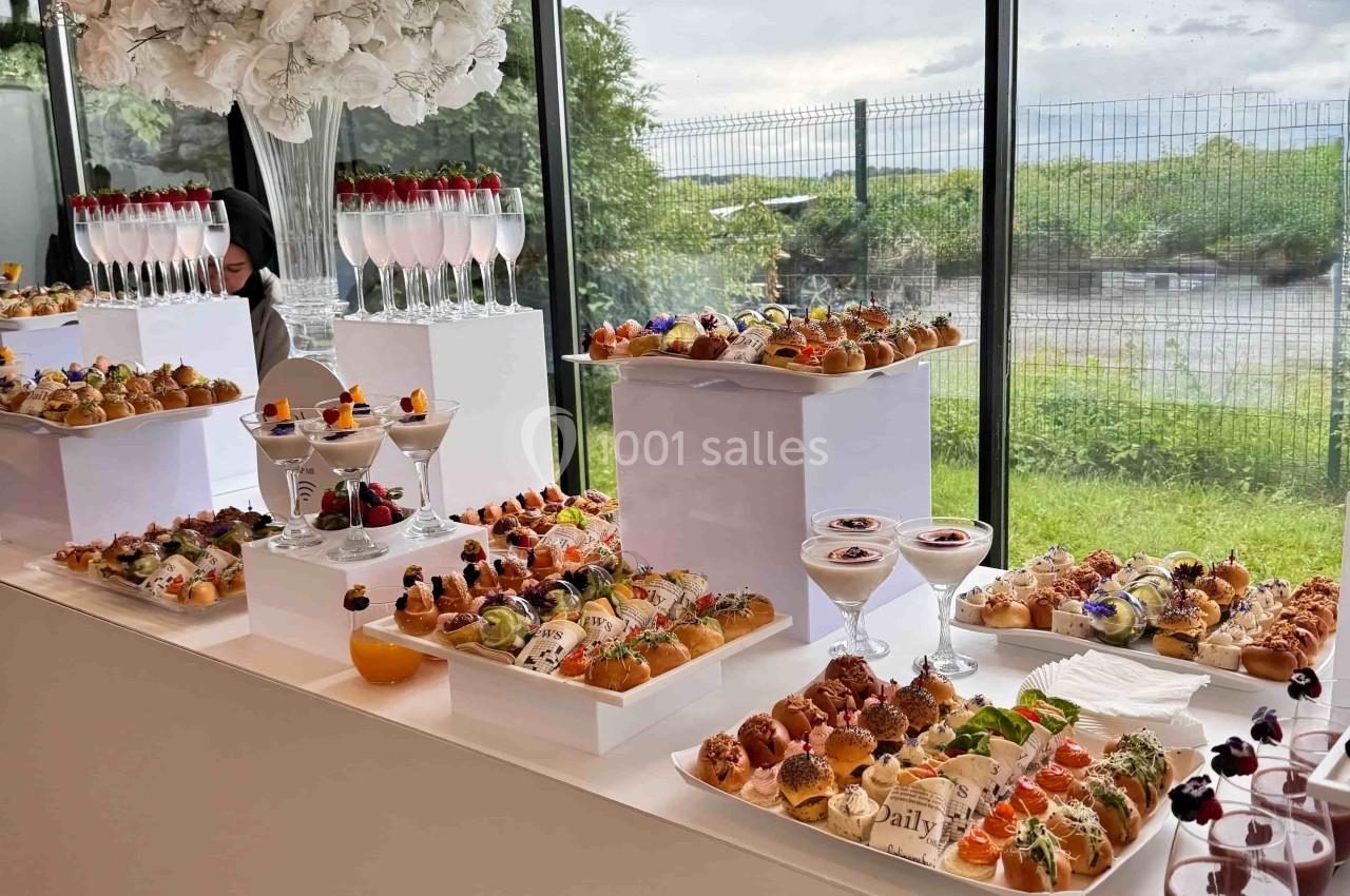 Buffet de pâtisseries et verrines élégamment disposé sur des tables blanches près d'une baie vitrée avec vue extérieure.