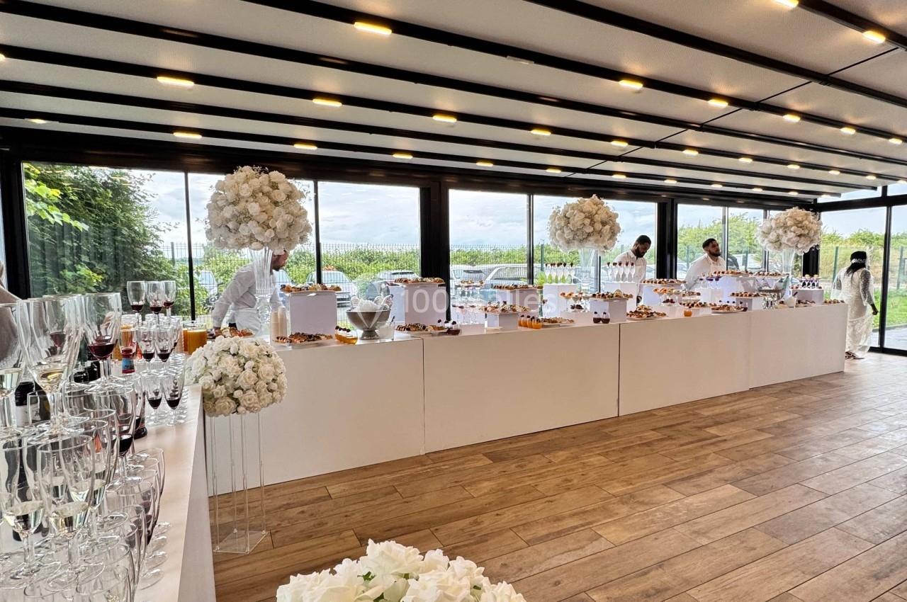 Salle lumineuse avec tables décorées de fleurs blanches, buffet varié et vue sur un paysage extérieur verdoyant.
