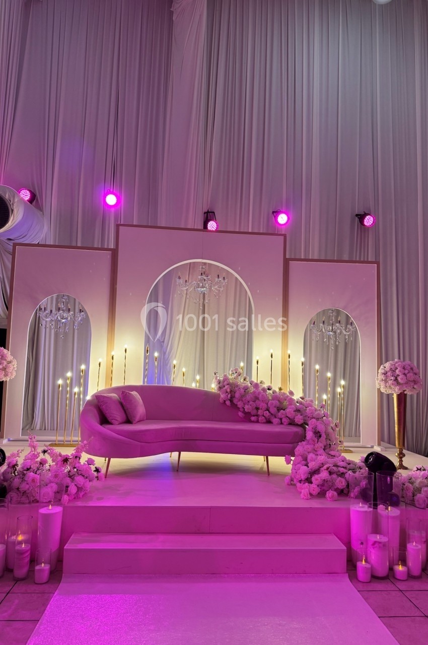 Décor de mariage élégant avec canapé rose, rideaux blancs, fleurs roses et éclairage violet tamisé.