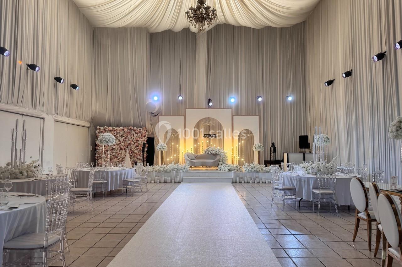 Salle de réception décorée avec des drapés blancs, des tables élégantes et une scène illuminée au fond.