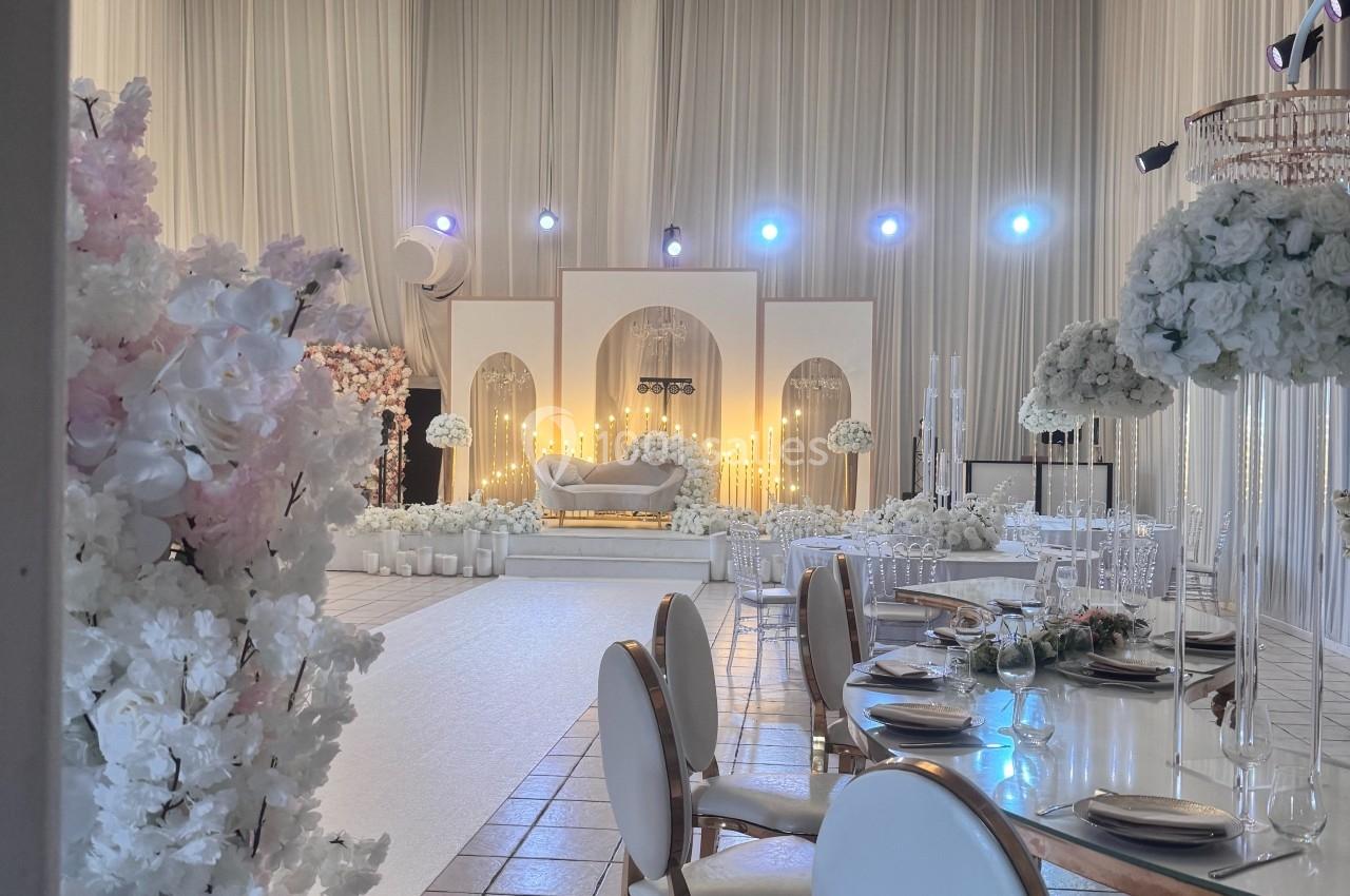 Salle de réception élégante avec décorations florales blanches, tables dressées et éclairage chaleureux.