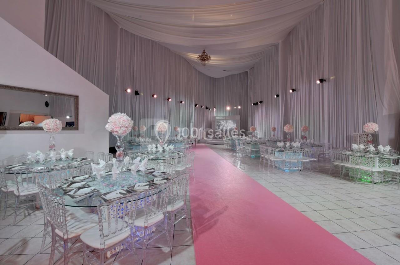 Salle de réception décorée avec des tables en verre, chaises transparentes, nappes blanches et allée centrale rose.