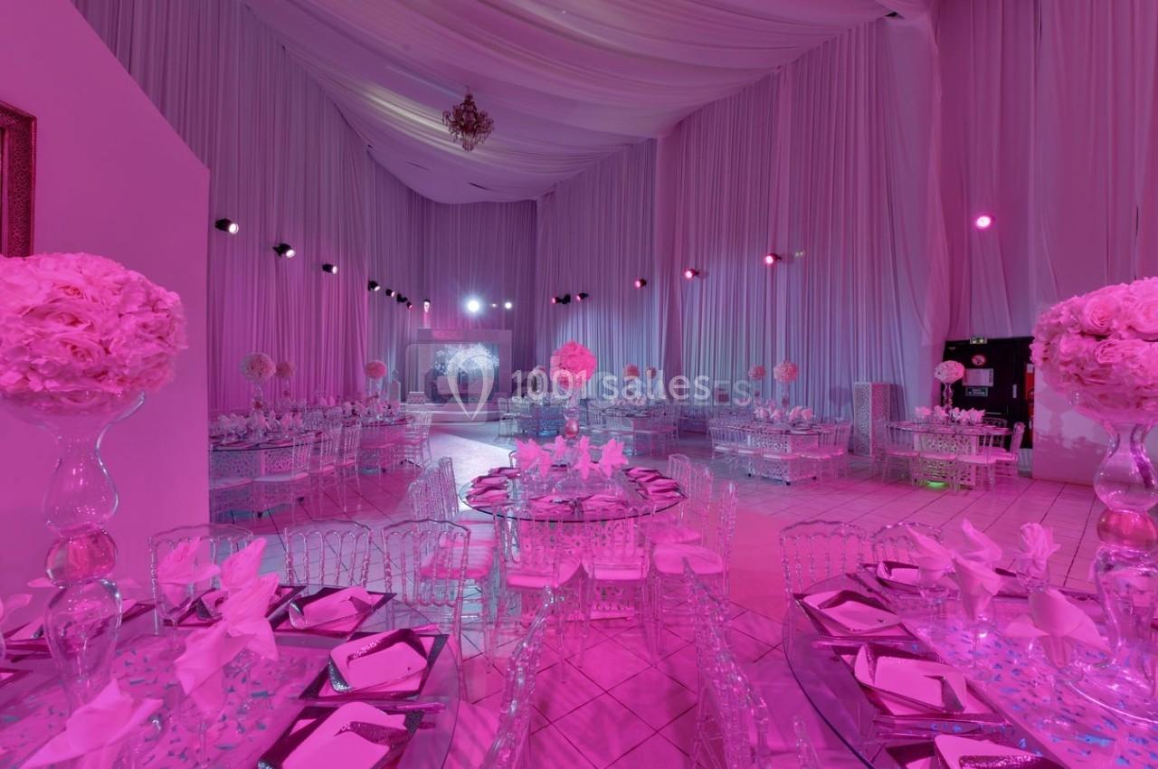 Salle de réception décorée en rose avec tables dressées, chaises transparentes et éclairage tamisé.