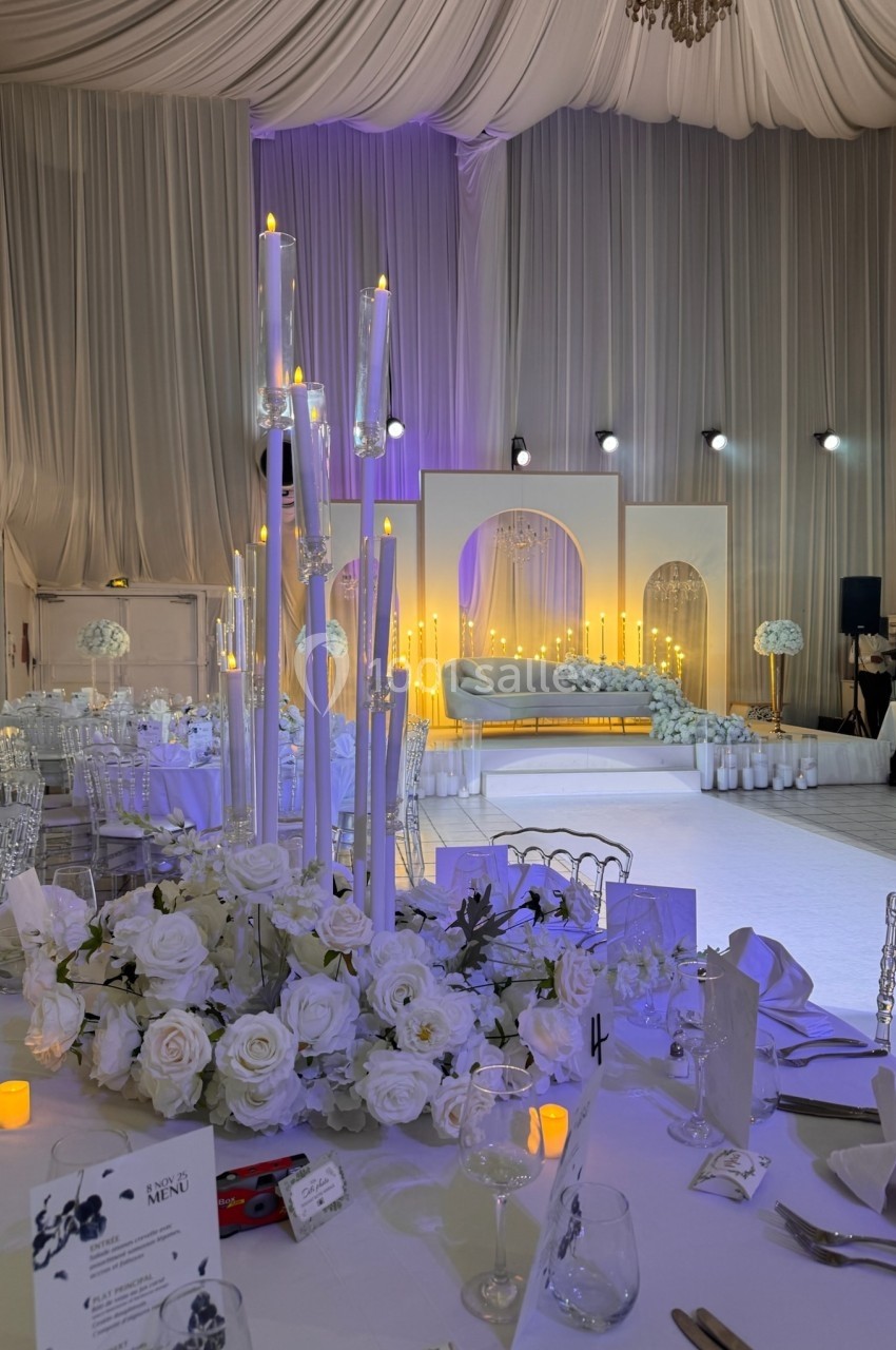 Salle de réception décorée avec des fleurs blanches, des chandeliers et une scène éclairée en arrière-plan.