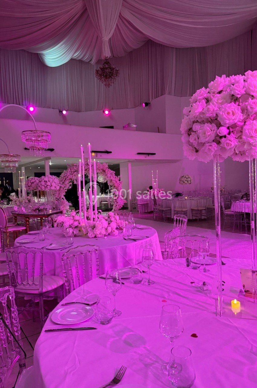 Salle de réception décorée de roses blanches, chandeliers et éclairage rose, avec tables dressées élégamment.