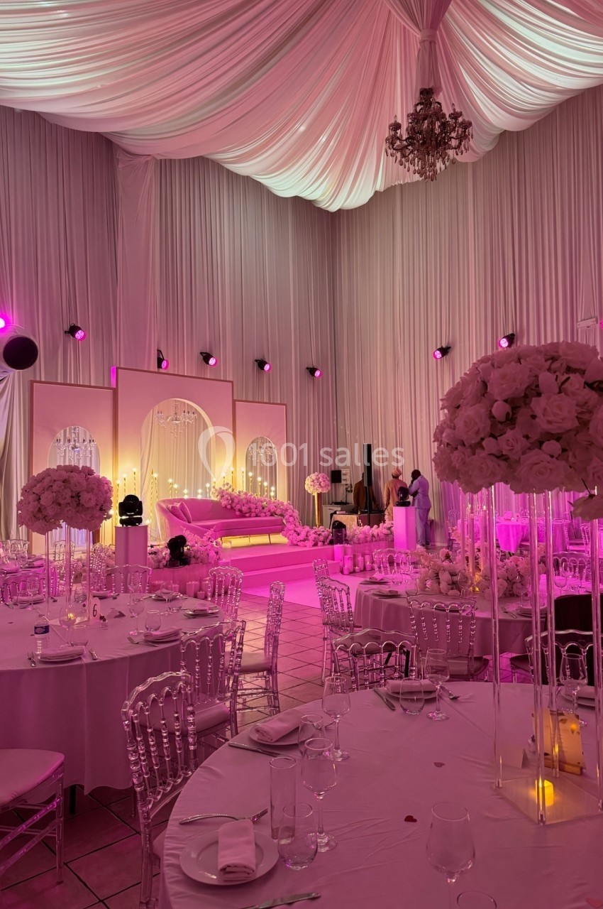 Salle de réception décorée avec des drapés blancs, éclairage rose, tables dressées et arrangements floraux élégants.