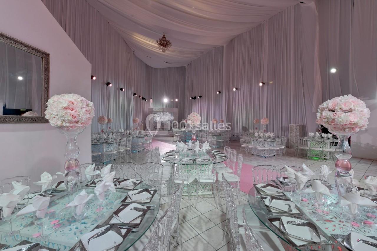Salle de réception décorée avec des tables en verre, des bouquets de fleurs roses et un plafond drapé de tissu blanc.
