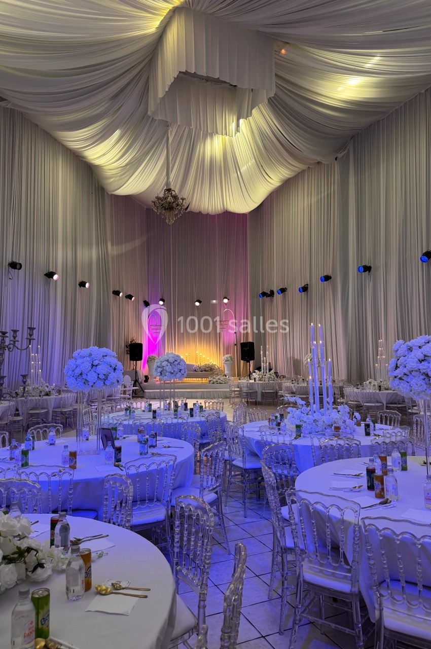 Salle de réception décorée avec des tables rondes, des chaises transparentes et des arrangements floraux blancs sous un…