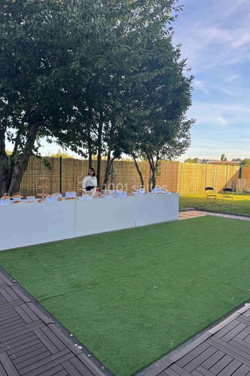 Table dressée en extérieur avec des amuse-bouches, située sur une pelouse artificielle près d'arbres et d'une clôture.