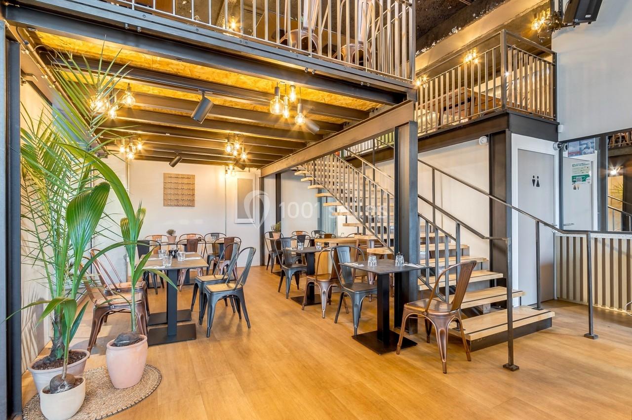 Salle de restaurant lumineuse avec des tables en bois, des chaises métalliques et une mezzanine accessible par un escalier.