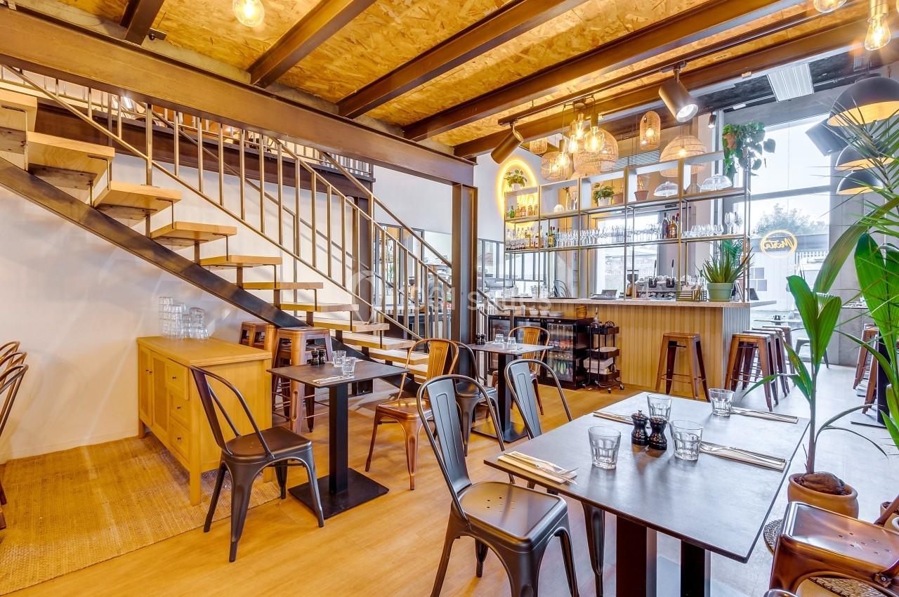 Salle de restaurant lumineuse avec mobilier en bois et métal, escalier en bois et bar en arrière-plan.