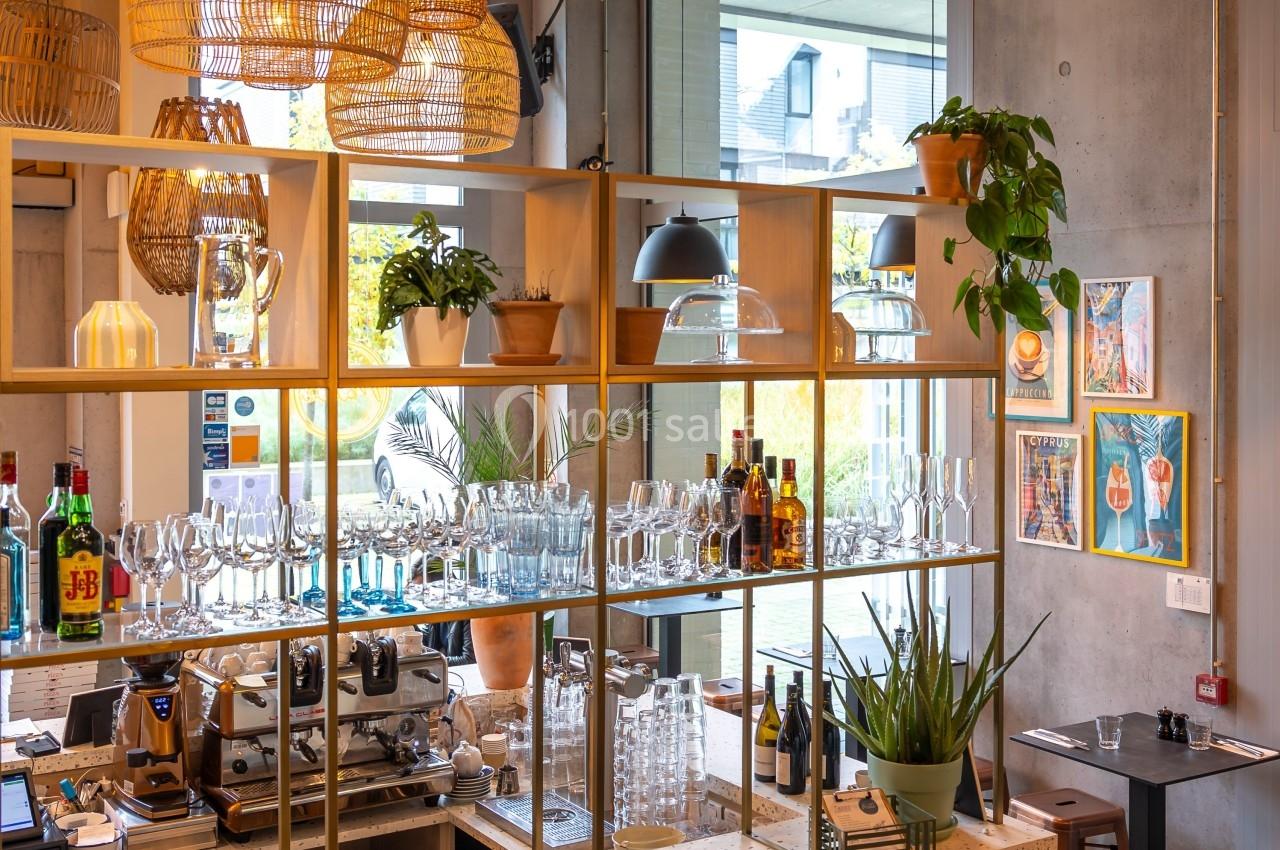 Étagères en bois avec verres, bouteilles et plantes dans un café lumineux au design moderne et chaleureux.
