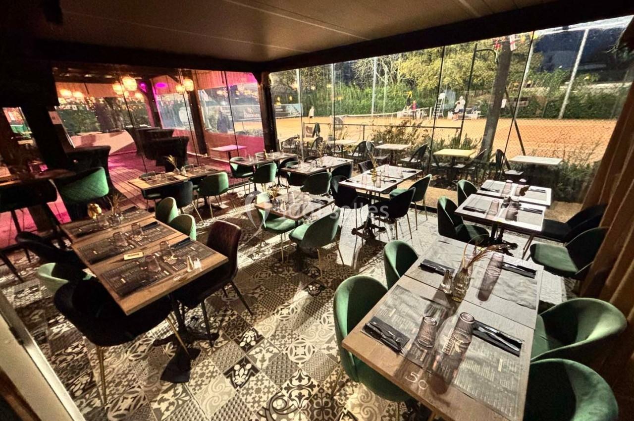 Salle de restaurant élégante avec tables dressées, chaises vertes et grandes baies vitrées donnant sur un extérieur éclairé.