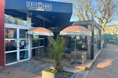 Miniature Location salle Le Bouscat (Gironde) - Côté Court #39 Entrée d'un café avec une grande terrasse, un parasol orange et des meubles en osier sous un ciel bleu.
