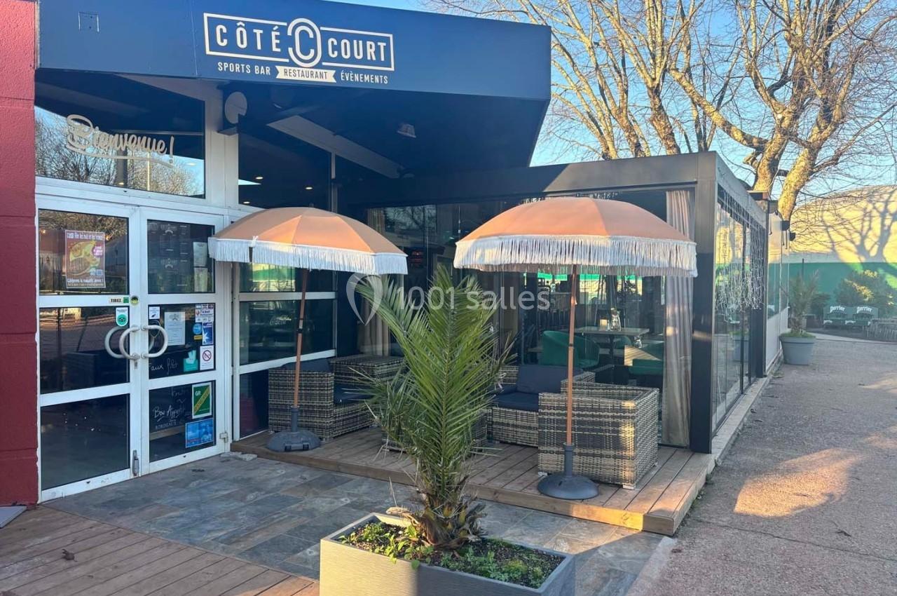 Entrée d'un restaurant avec terrasse aménagée, parasols, mobilier en rotin et plantes, par temps ensoleillé.
