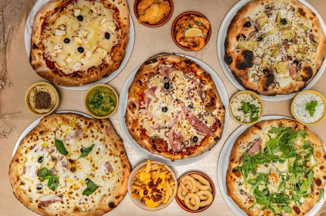 Assortiment de pizzas variées avec garnitures différentes, entourées de petits plats d'accompagnement sur une table en bois.