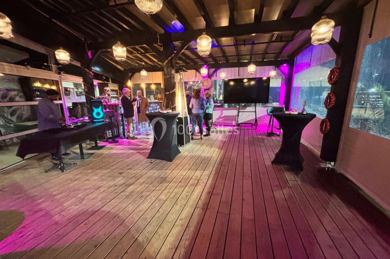 Salle en bois avec éclairage tamisé, tables hautes, écran, DJ et quelques personnes debout près d'une scène.