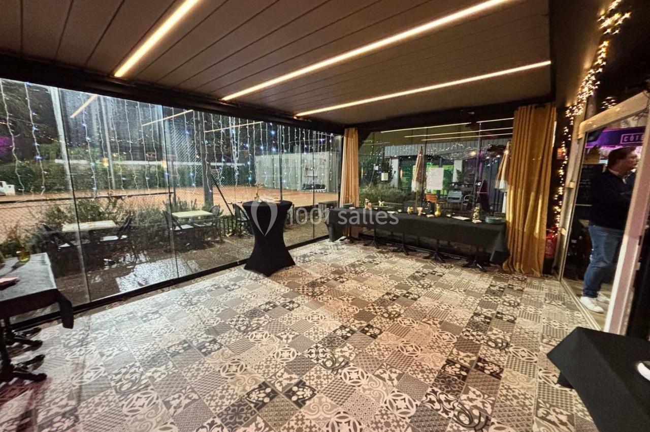 Salle avec sol en carreaux de ciment, grandes baies vitrées donnant sur un extérieur éclairé et tables dressées.