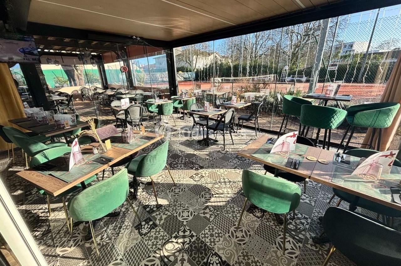 Terrasse couverte d'un restaurant avec mobilier vert et noir, sol en carreaux décoratifs, vue sur des terrains de sport.