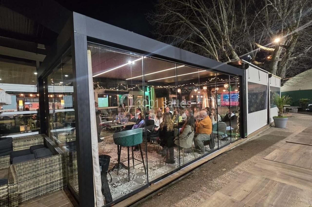Groupe de personnes assises à l'intérieur d'une terrasse vitrée éclairée, entourée d'arbres en soirée.