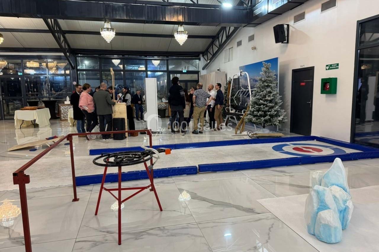 Des personnes discutent près d'une piste de curling intérieure dans une salle lumineuse avec décor hivernal.