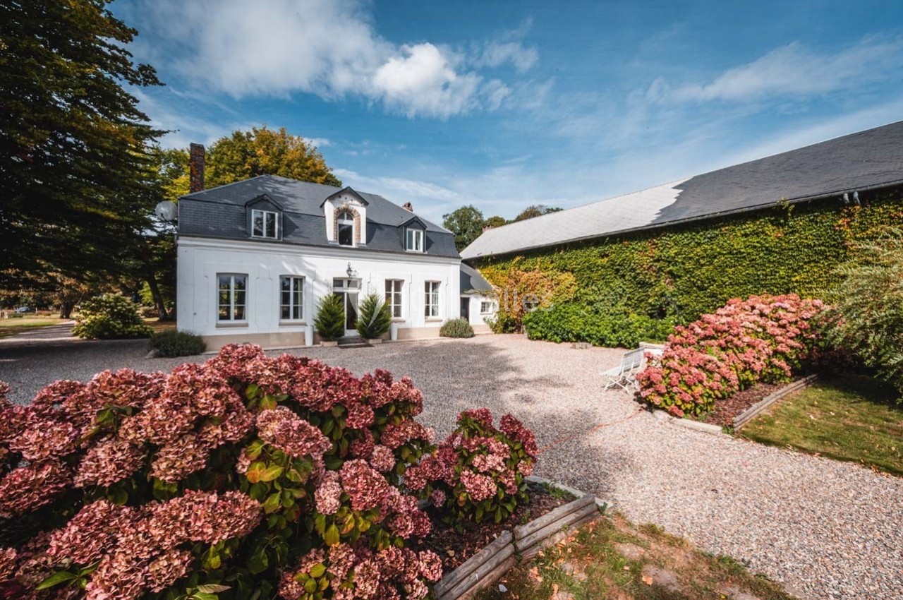 Maison blanche avec toit en ardoise, entourée d'hortensias et d'une cour gravillonnée, sous un ciel bleu.