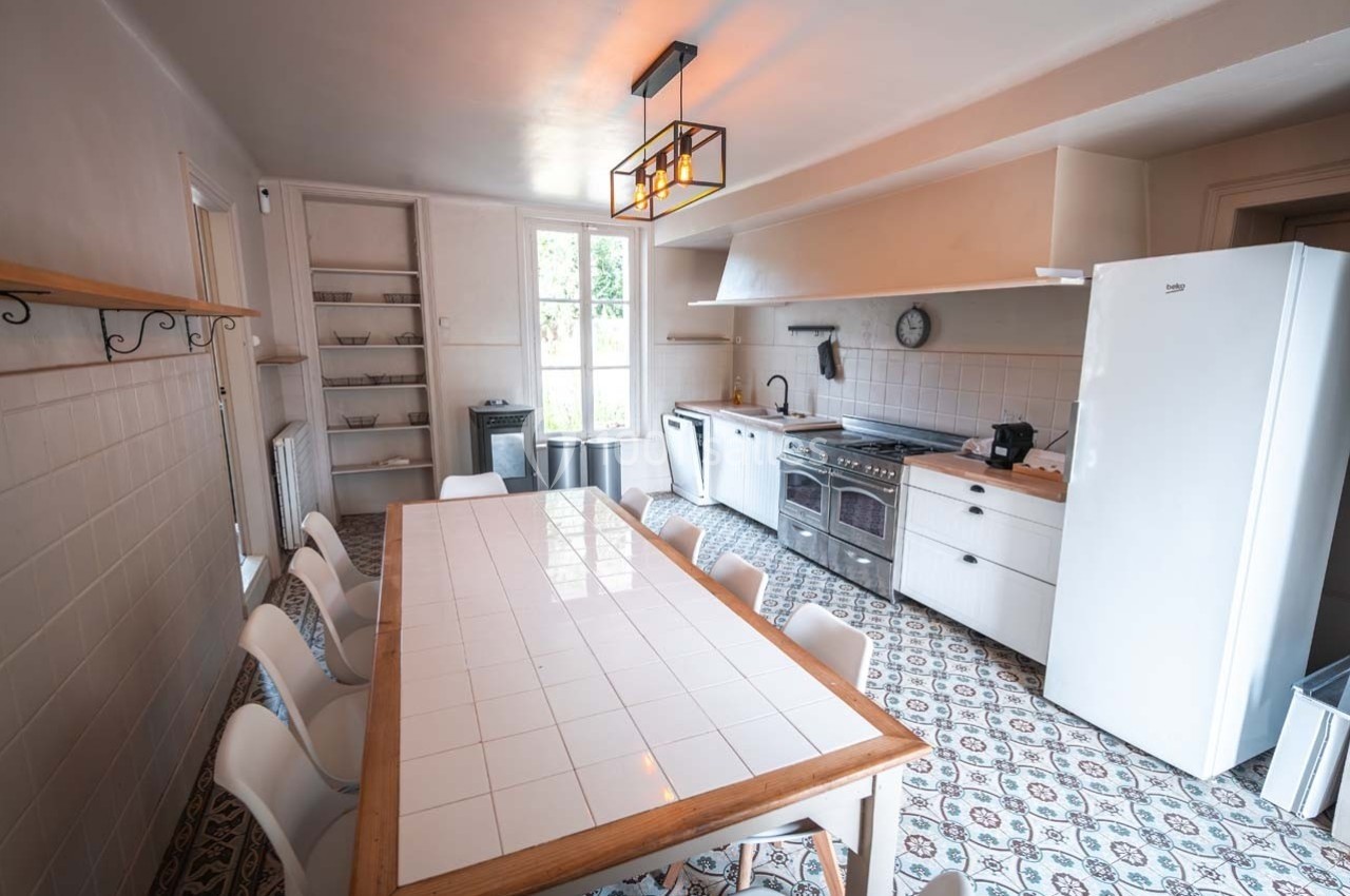 Cuisine lumineuse avec une grande table en bois, des chaises blanches, un réfrigérateur, une cuisinière et des étagères…