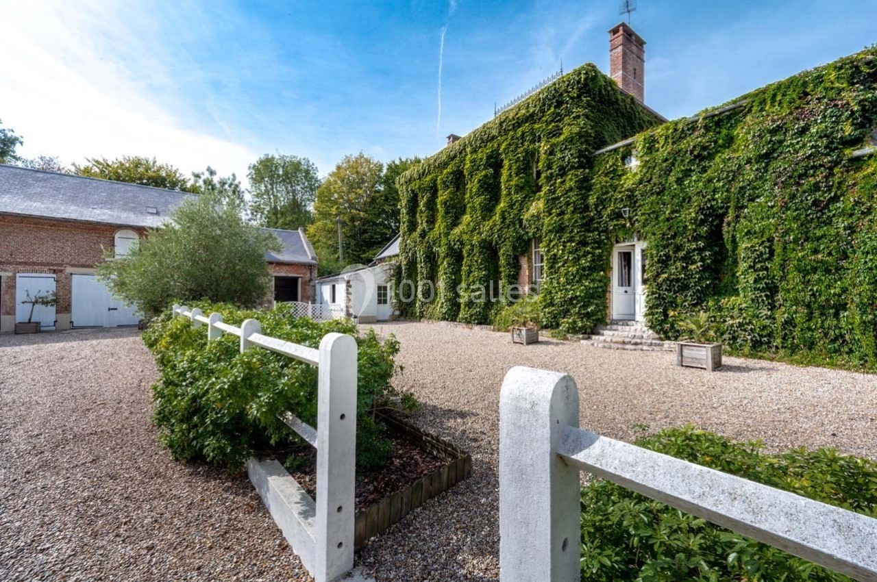 Cour pavée bordée de bâtiments en briques et d'une maison recouverte de vigne, sous un ciel bleu dégagé.
