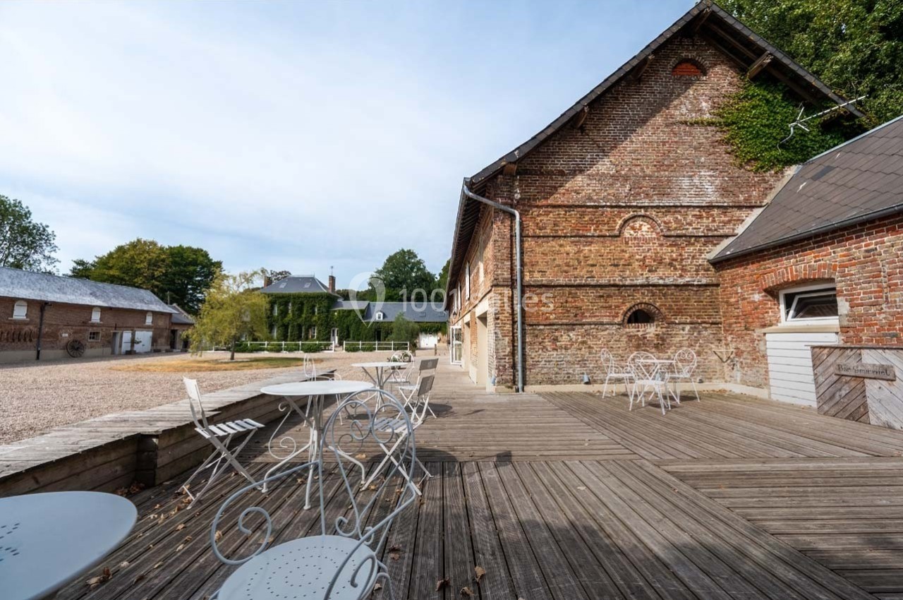 Terrasse en bois avec tables et chaises blanches, entourée de bâtiments en briques et végétation en arrière-plan.