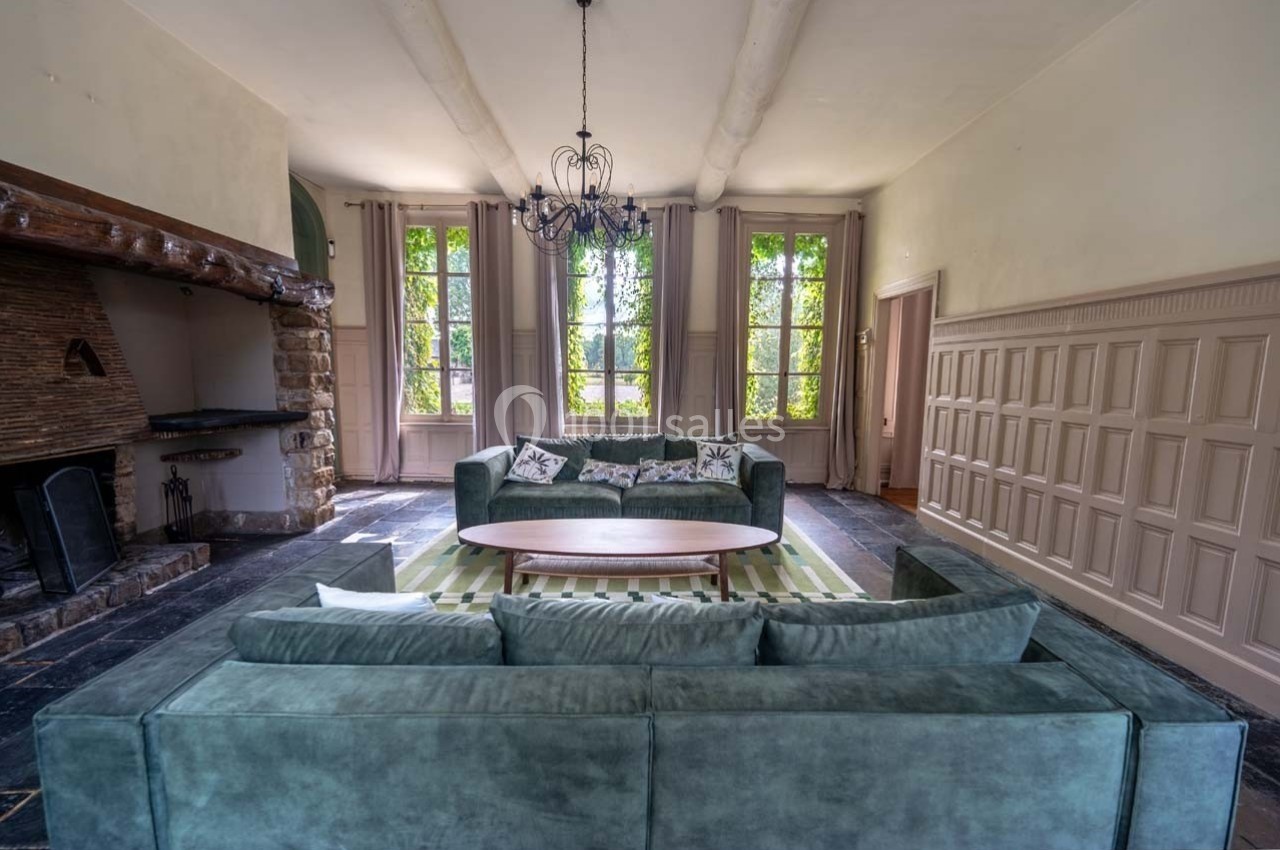 Salon lumineux avec canapé vert, table basse en bois, cheminée en pierre et grandes fenêtres donnant sur un jardin.