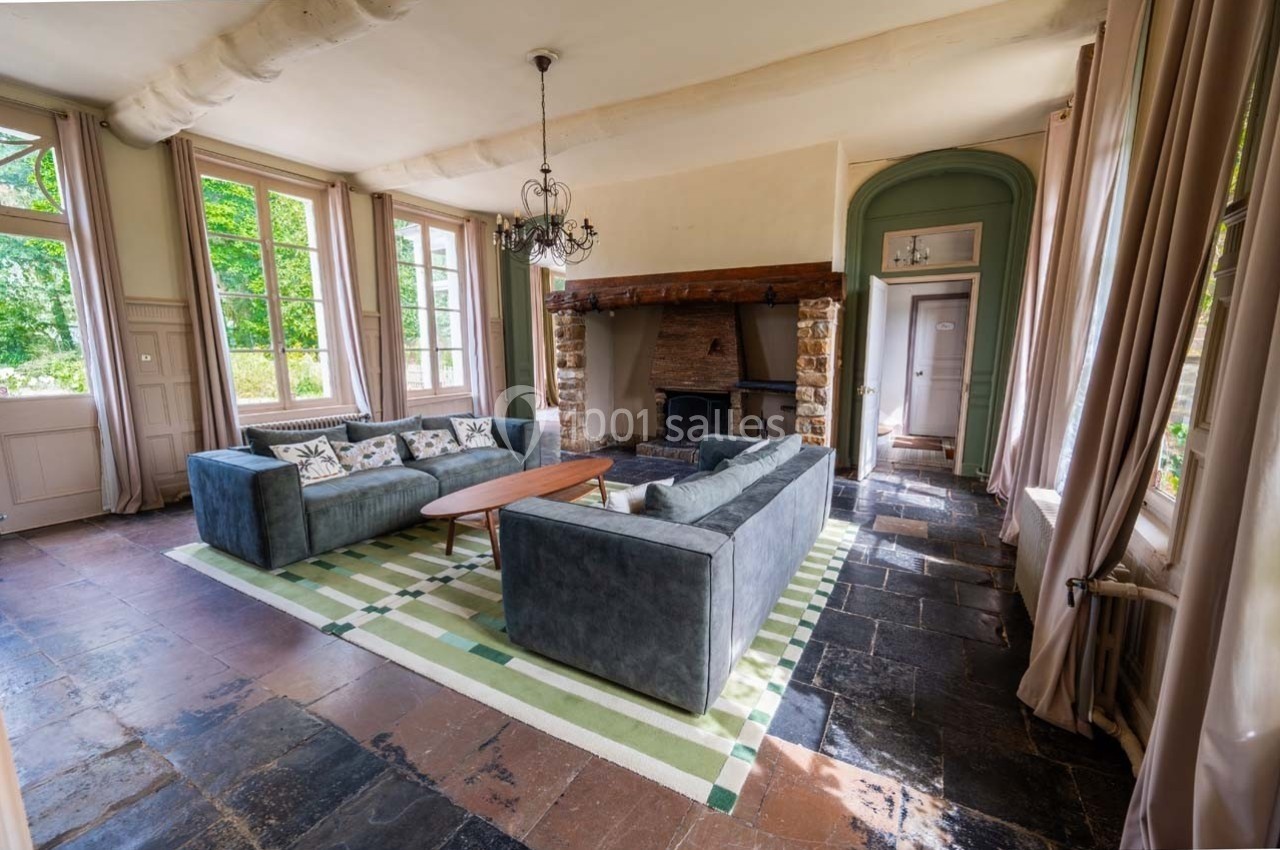 Salon lumineux avec canapés gris, cheminée en pierre, tapis vert et grandes fenêtres donnant sur un jardin verdoyant.