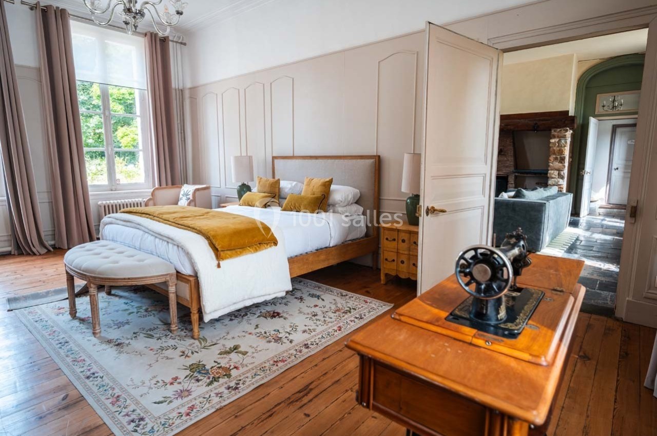 Chambre lumineuse avec lit double, coussins jaunes, tapis floral, machine à coudre ancienne et vue sur un salon en arrière…