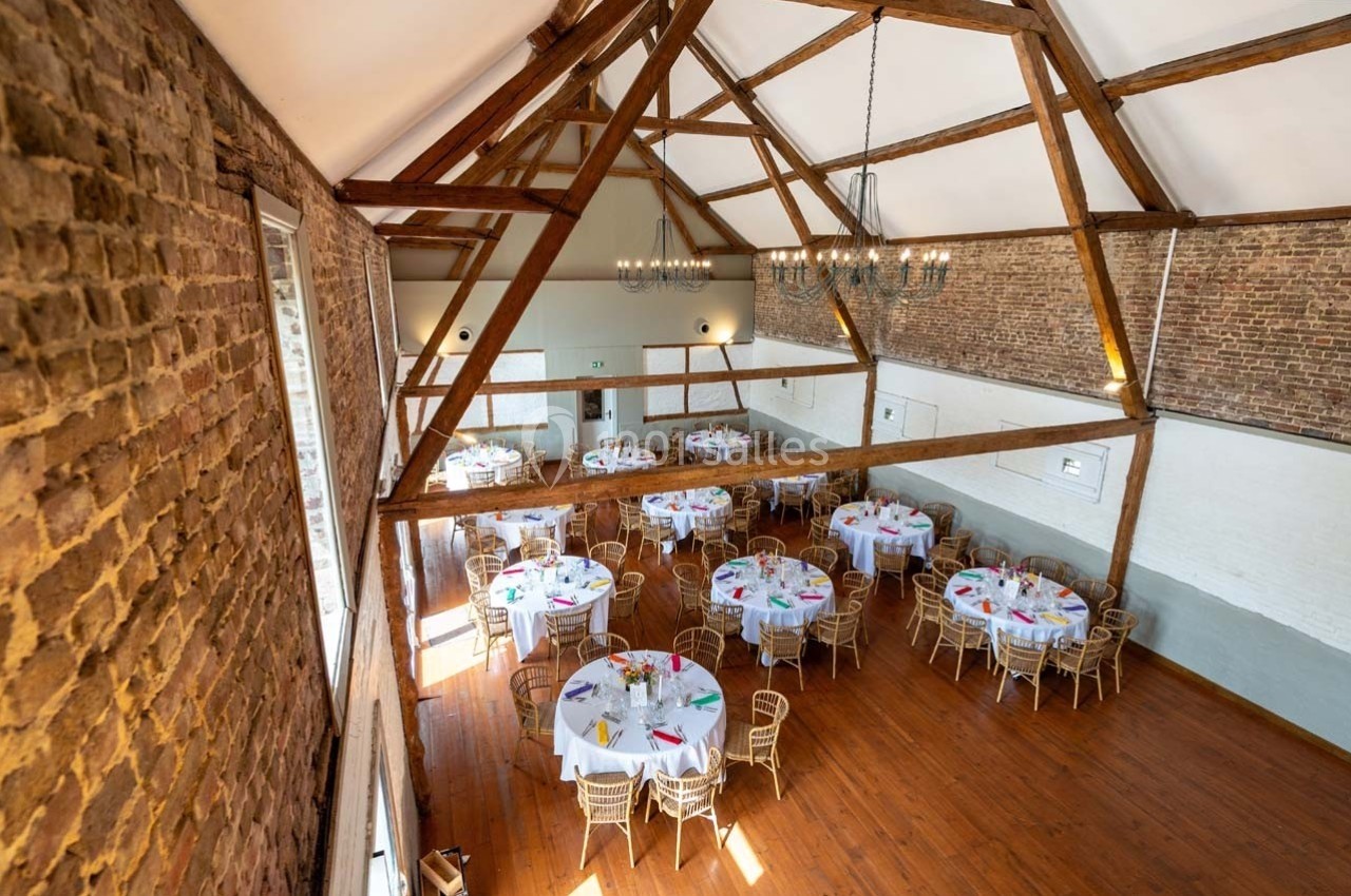 Salle de réception avec murs en briques, poutres apparentes et tables rondes dressées pour un événement.