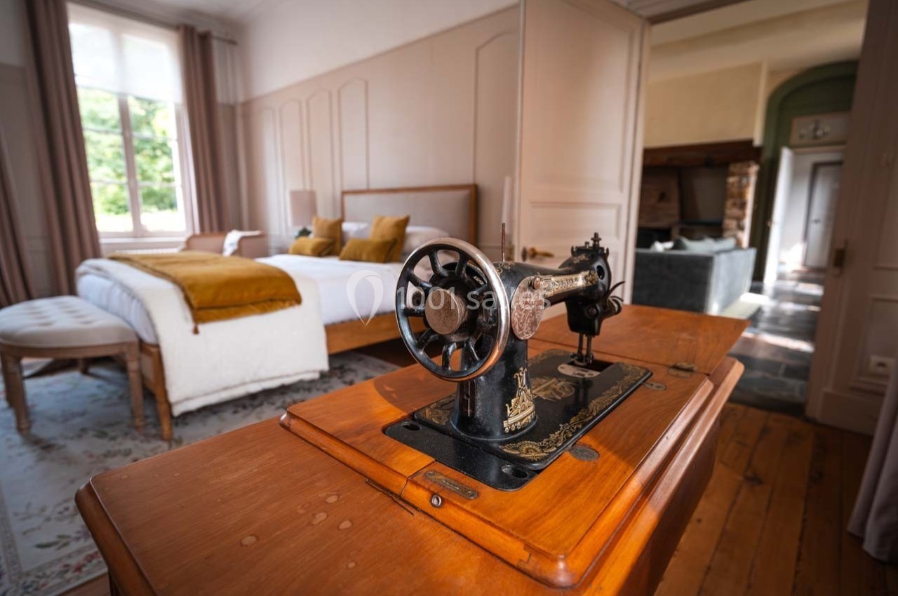 Machine à coudre ancienne en bois et métal dans une chambre lumineuse avec lit double et décor chaleureux en arrière-plan.