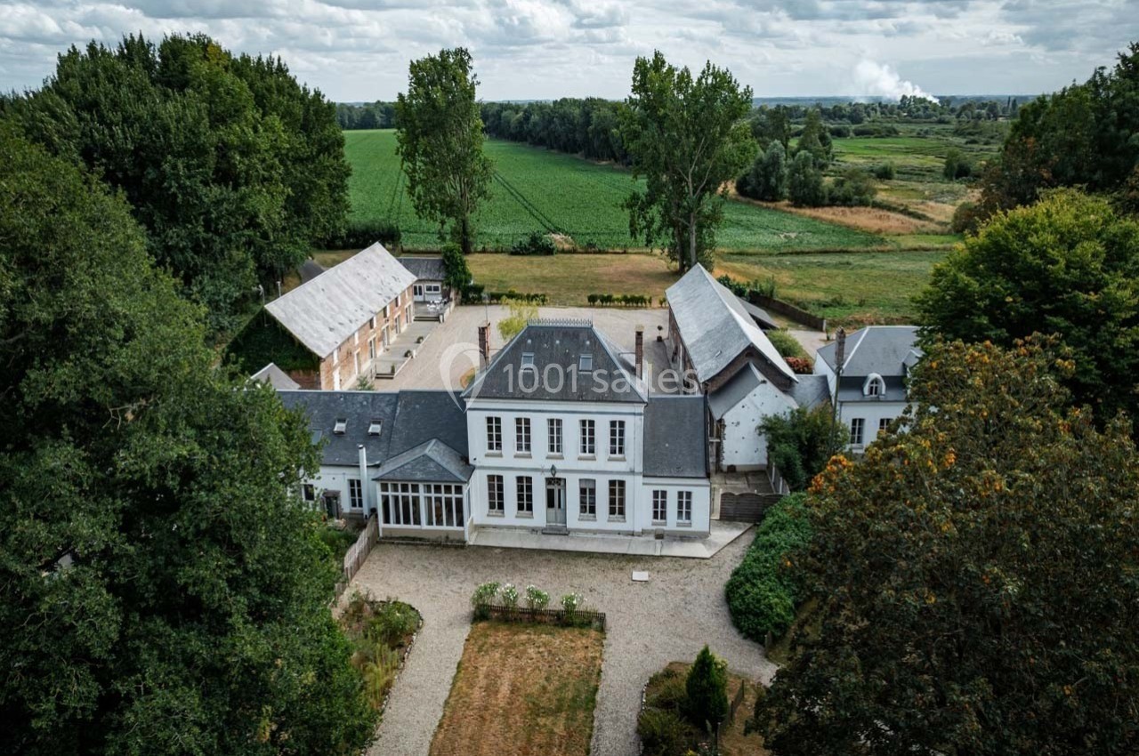 Vue aérienne d'une grande maison blanche entourée de jardins, d'arbres et de bâtiments annexes en pleine campagne.