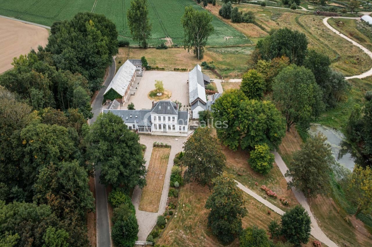Vue aérienne d'un domaine rural avec bâtiments entourés de jardins, d'arbres et de champs verdoyants.
