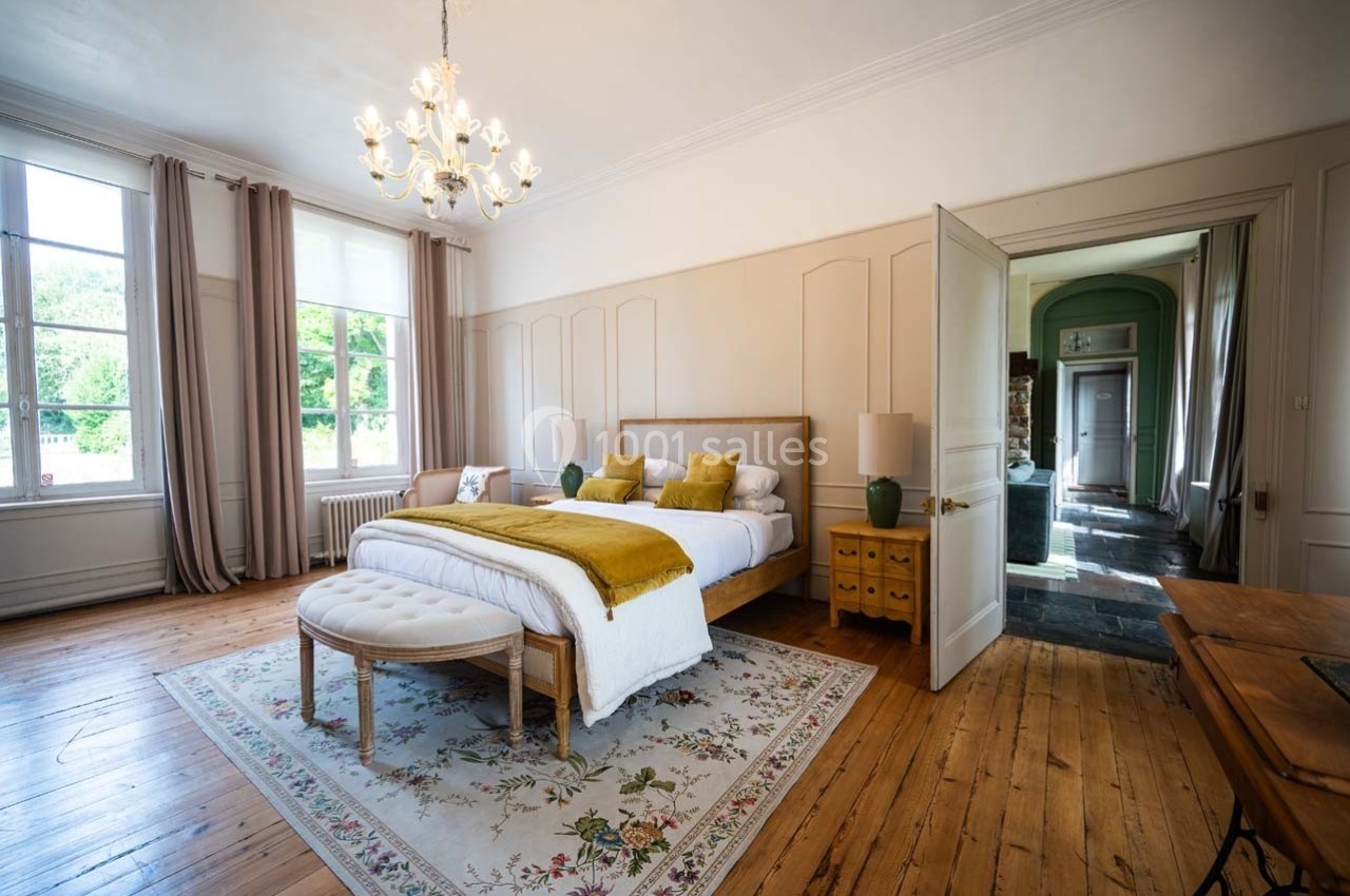 Chambre lumineuse avec lit double, couvre-lit jaune, tapis floral, parquet en bois et grandes fenêtres avec rideaux.