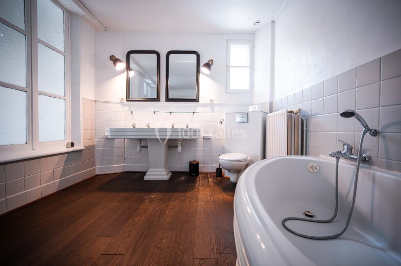 Salle de bain lumineuse avec baignoire, double vasque, carrelage blanc, parquet foncé et fenêtres vitrées.