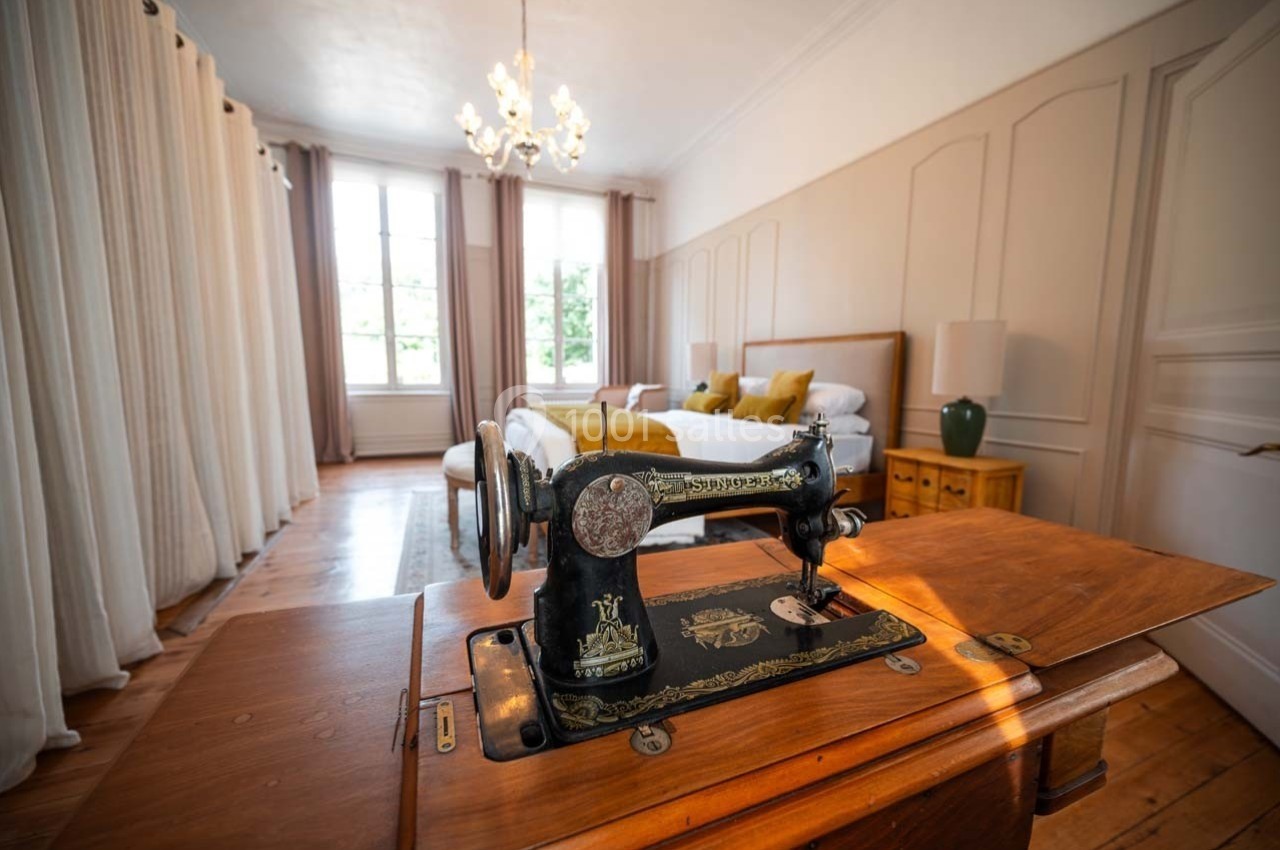 Machine à coudre ancienne en bois au premier plan dans une chambre lumineuse avec lit, rideaux et lustre.