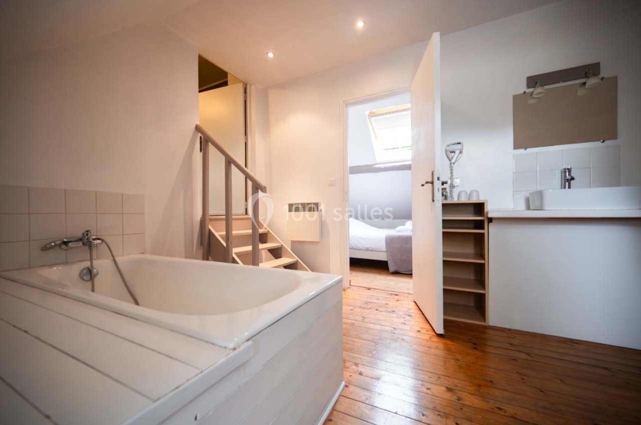 Salle de bain lumineuse avec baignoire, meuble vasque, escalier en bois et vue sur une chambre adjacente.