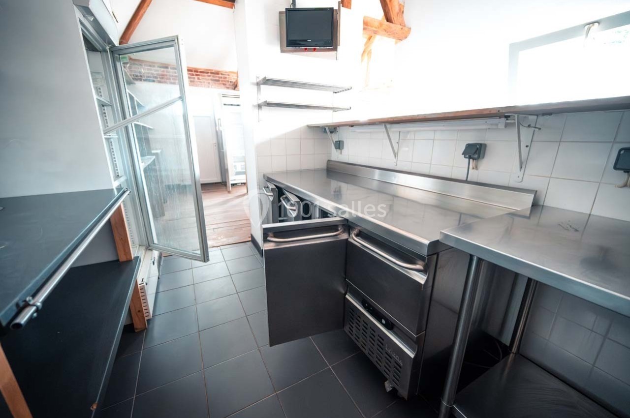 Cuisine professionnelle avec plans de travail en inox, étagères murales, réfrigérateur ouvert et carrelage sombre.
