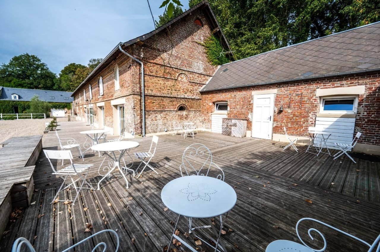 Terrasse en bois avec tables et chaises blanches devant un bâtiment en briques, entourée de verdure.