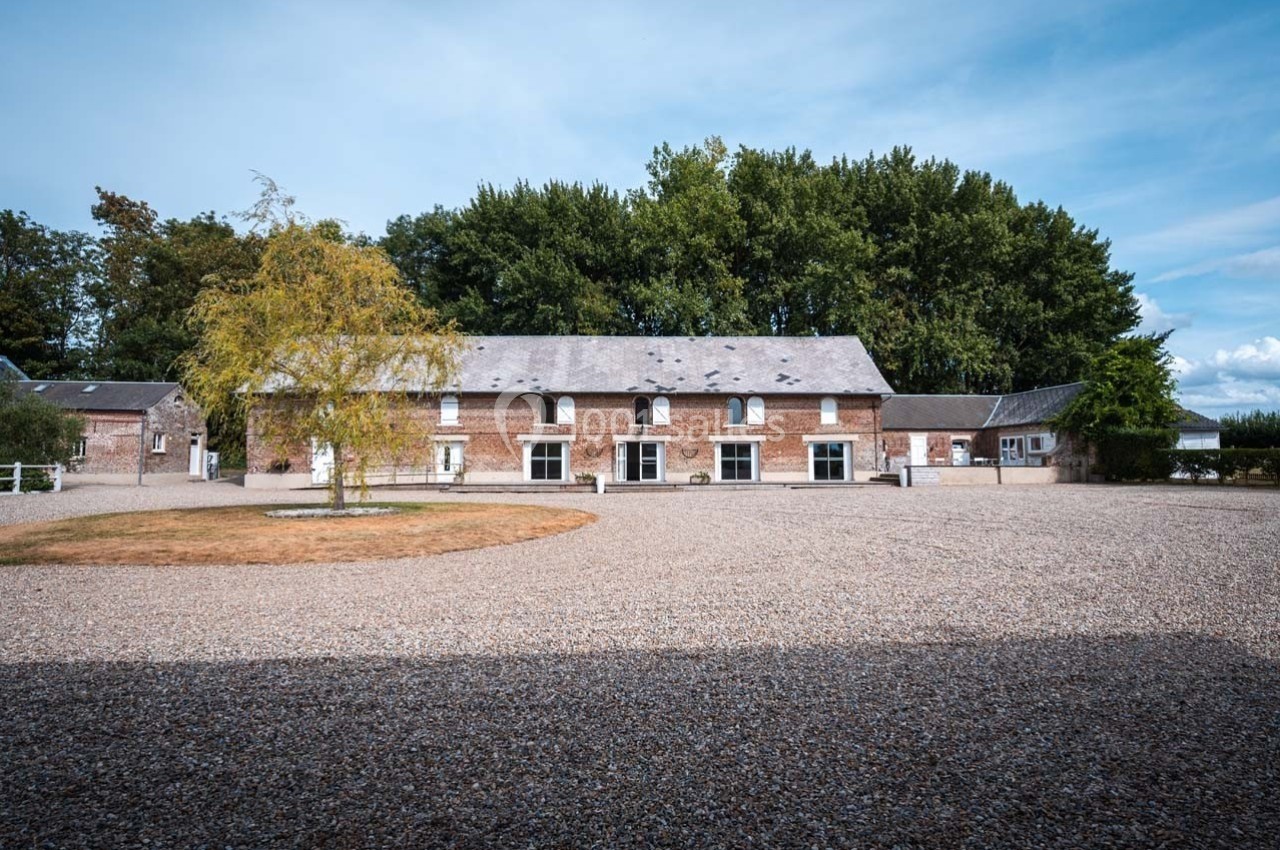 Bâtiment en briques avec plusieurs fenêtres, entouré d'une cour en gravier et d'arbres en arrière-plan.
